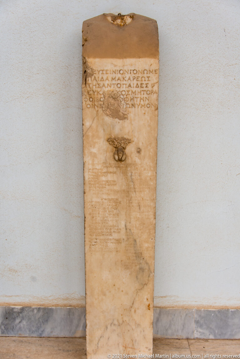 Tall narrow marble stele with Greek text at top, dense smaller lines below, and metal mounting hardware on the face.