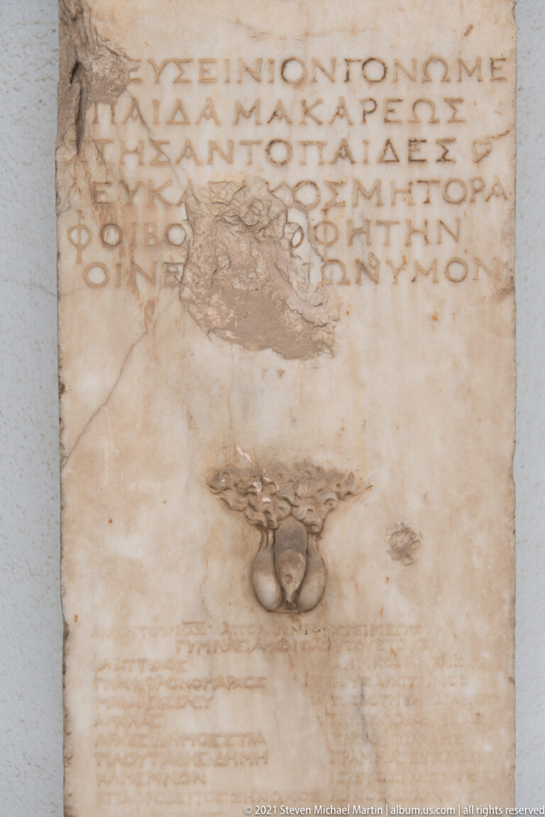 Close-up of Greek capital letters on a marble stele above a dark metal mounting fitting.