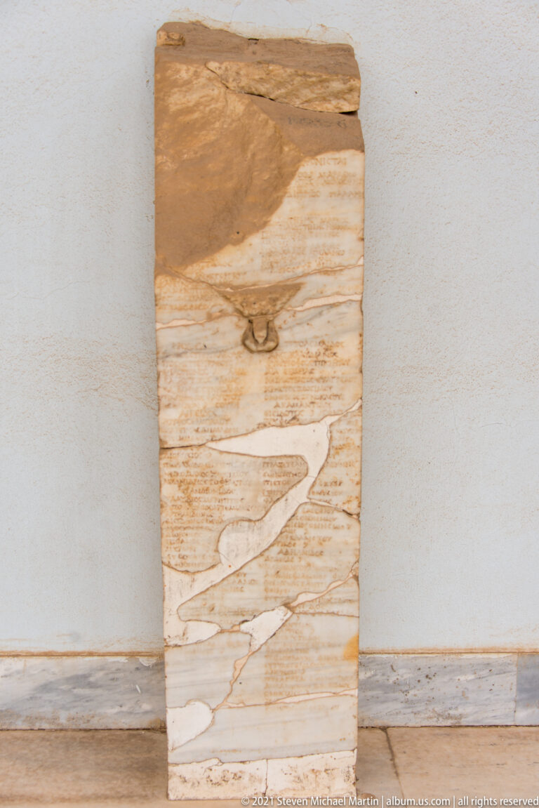 Reverse of a tall pale marble stele with strong veining, an irregular top, and a small metal fitting.
