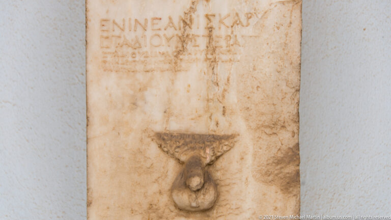 Close view of a narrow marble stele with several lines of Greek letters and a dark ring-shaped metal fitting.