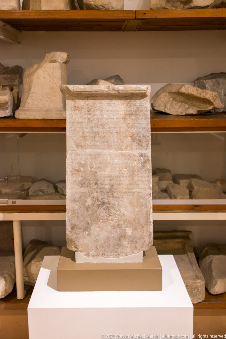 Large rectangular pale stone block with faint inscription on a pedestal, with shelves of stone fragments behind it.