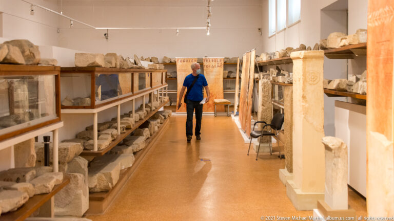 Visitor walking through a narrow museum aisle lined with shelves and many stone inscription fragments.
