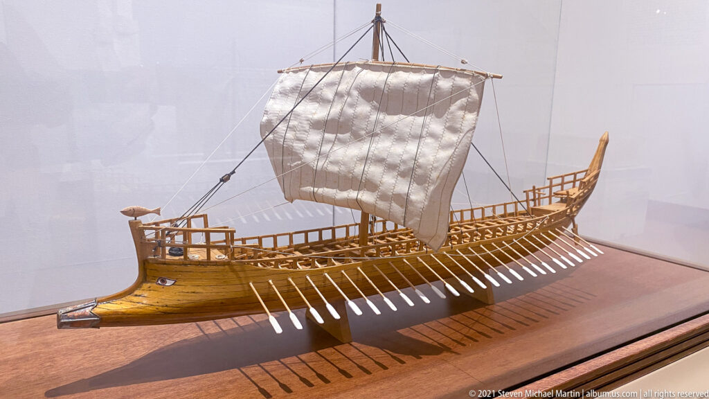 Detailed wooden model of an ancient oared ship with a square sail, many oars, and raised bow and stern.