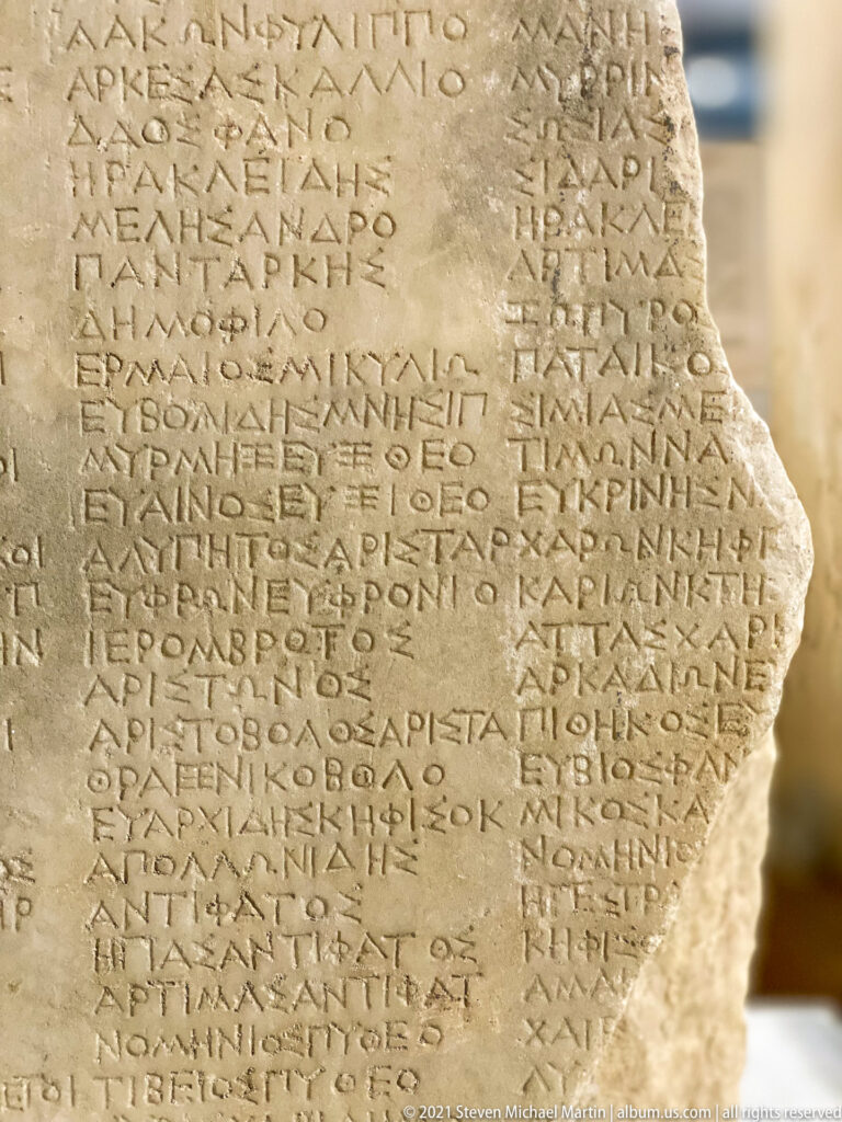 Broken pale marble slab densely carved with rows of Greek capital letters, with an irregular missing edge at right.