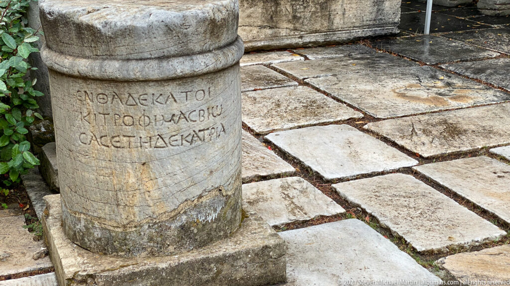 Short cylindrical stone monument beside paving, with several lines of Greek letters carved around its shaft.