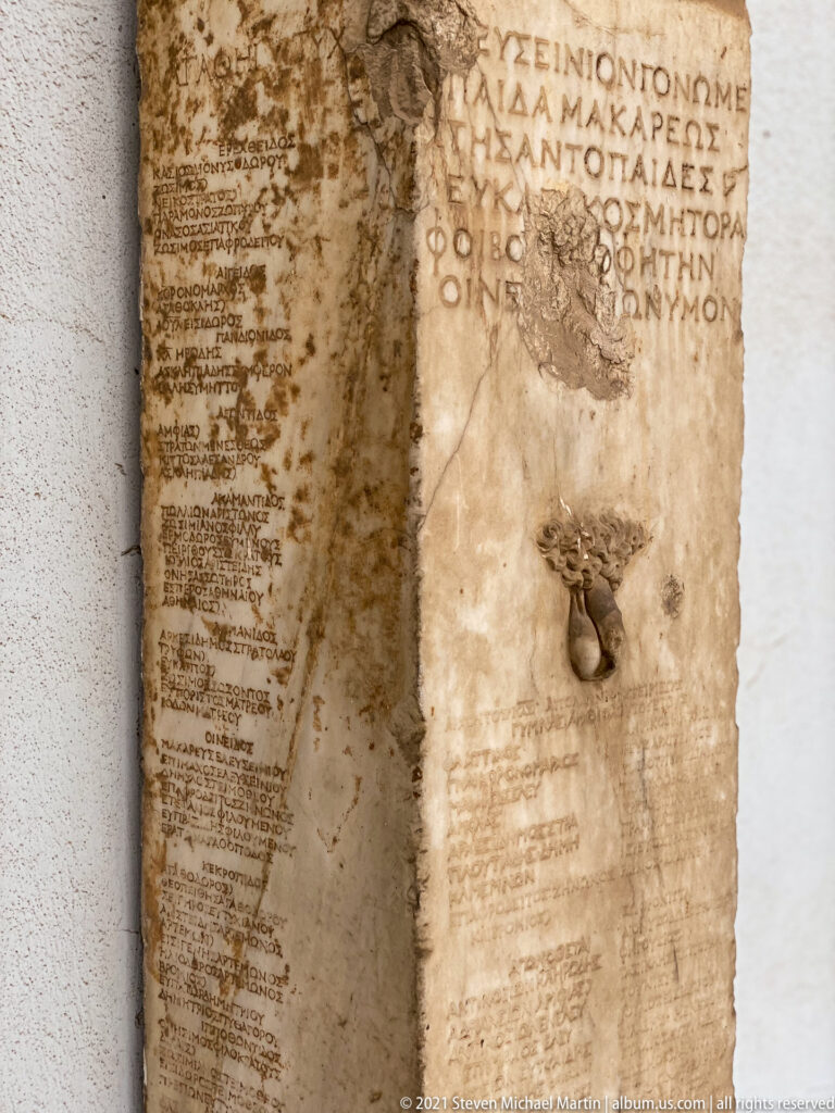 Tall narrow marble stele with large Greek letters above dense smaller text and dark metal fittings.