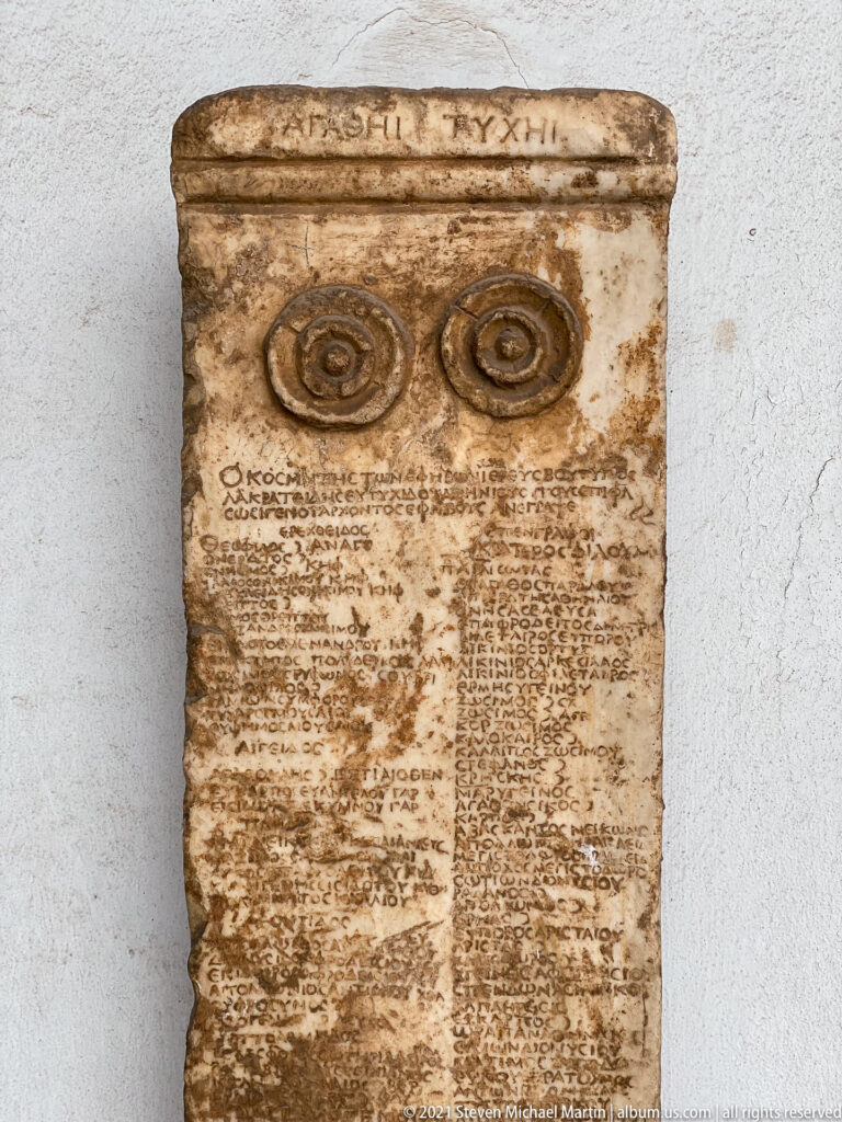 Tall marble stele covered with Greek lettering and two circular carved ornaments near its upper section.