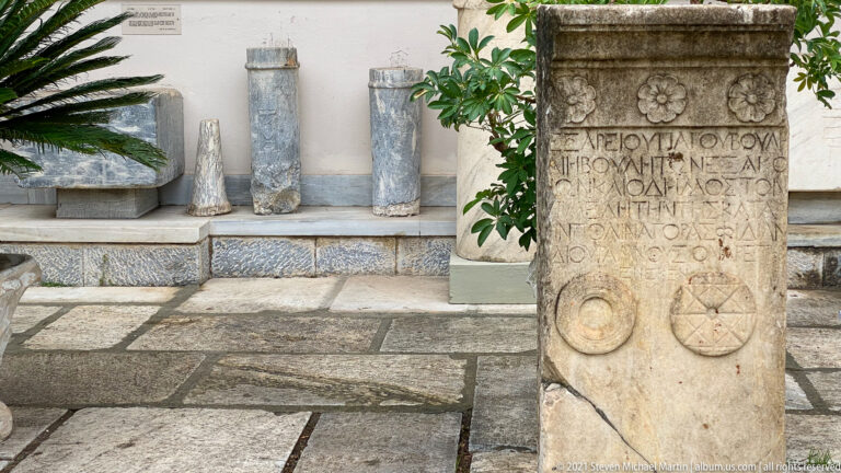 Upright marble stele with Greek inscription, floral rosettes across the top, and two circular carved symbols below.