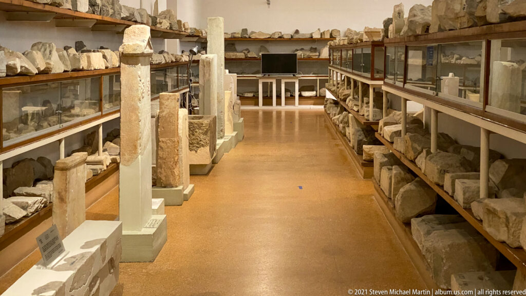 Long museum aisle with upright inscribed stones in the center and shelves of stone fragments along both walls.