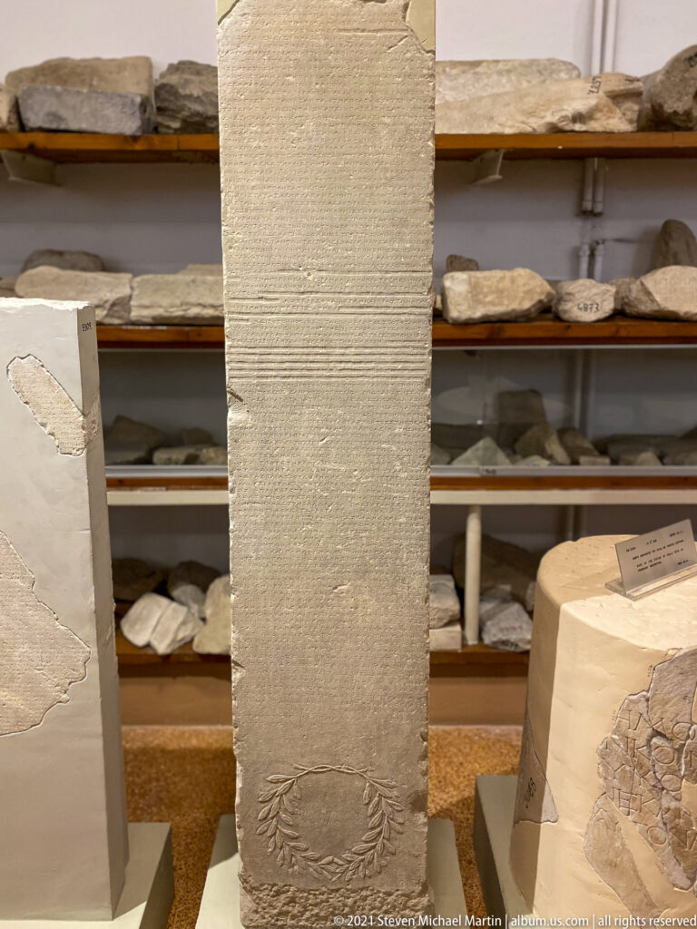 Tall narrow marble stele with dense Greek inscription and a carved wreath near the lower edge.