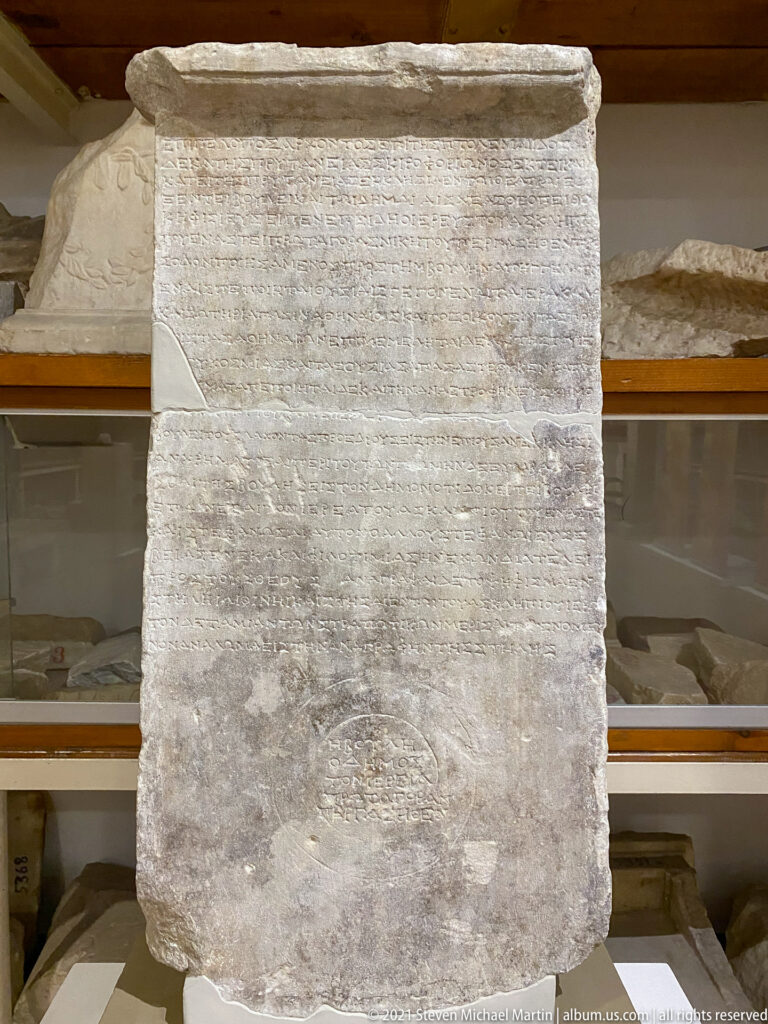 Weathered rectangular marble stele with faint Greek lettering, projecting top molding, and heavily abraded lower surface.