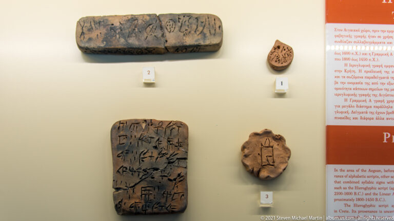 Four small brown clay objects with incised signs mounted on a museum wall beside an explanatory panel.
