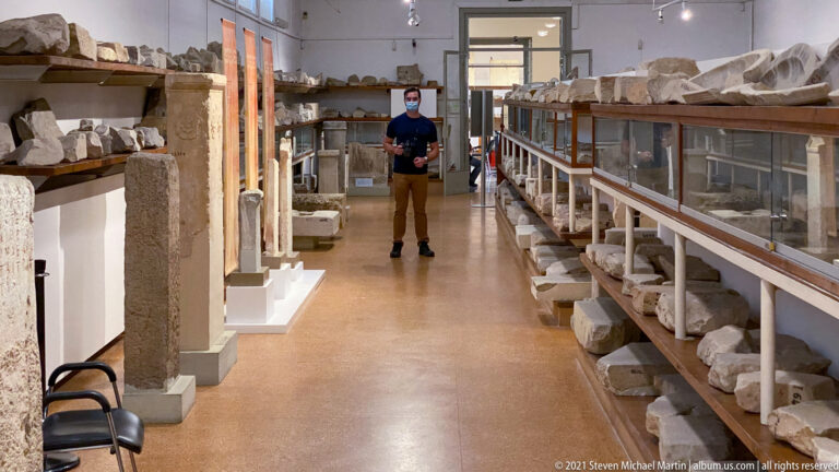 Masked visitor standing in a long museum aisle lined with upright stelae, shelves, and stone fragments.