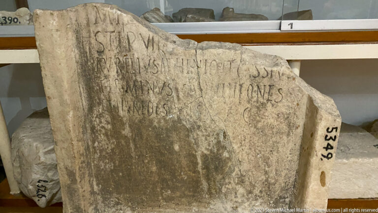 Irregular stone fragment with several lines of large weathered capital letters and broken edges.