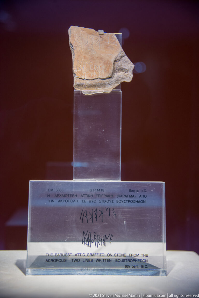 Small pale stone fragment above a transparent panel reproducing two lines of ancient Greek letters.