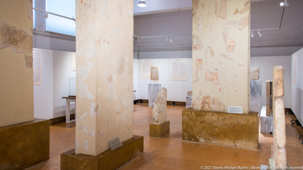 Museum gallery with tall piers and numerous inscribed stone slabs and stelae displayed around the room.
