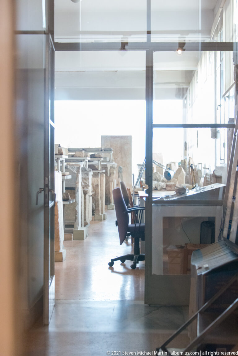 View through glass into a museum work area containing many stone inscriptions, tables, shelving, and an office chair.