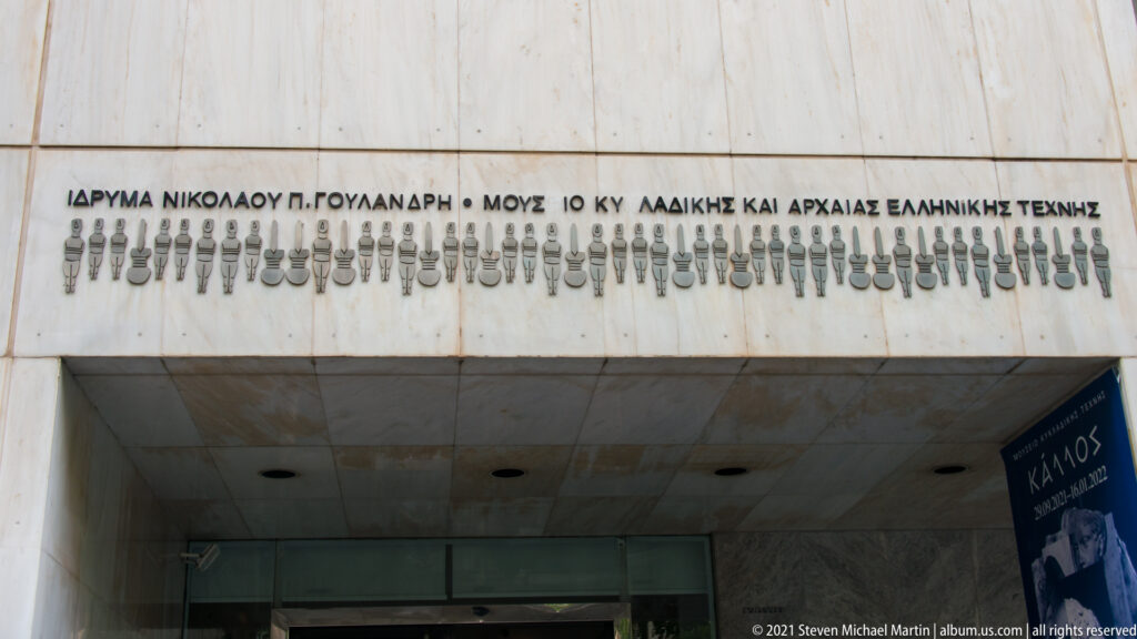 Marble museum entrance with Greek inscription, stylized figure motif, recessed glass doors, and an exhibition banner at right.