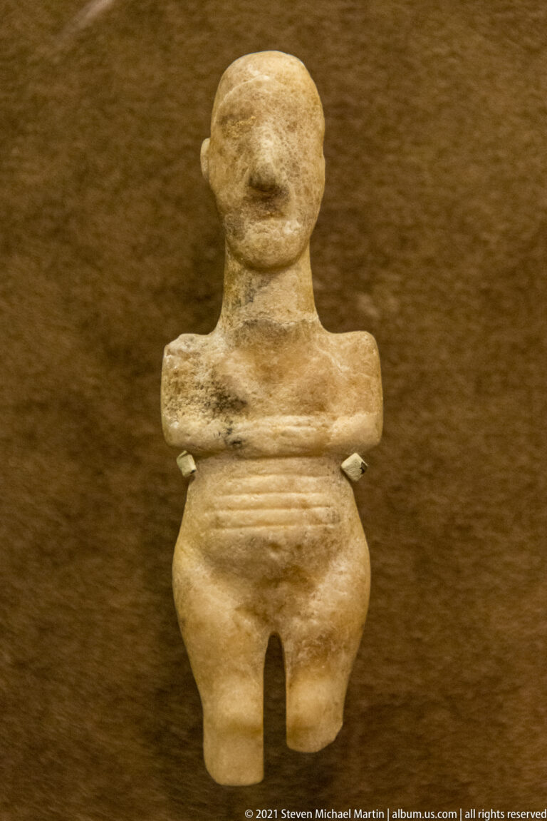 Stocky pale marble female figurine with rounded head, folded arms, broad abdomen, and separated legs.