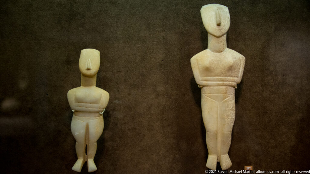 Two pale marble Cycladic folded-arm female figurines, one small and compact and one tall and slender, displayed together.