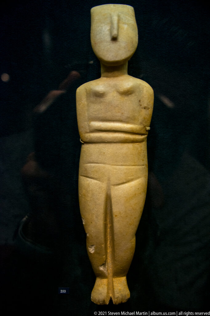Broad pale marble Cycladic female figure with flattened head, projecting nose, folded arms, and deeply divided legs.