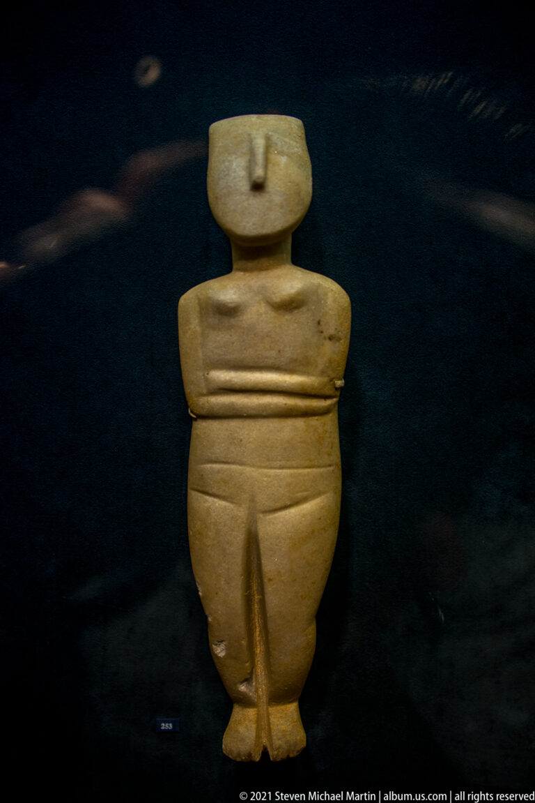 Frontal view of a broad Cycladic marble female figurine with folded arms, rounded abdomen, and separated legs.