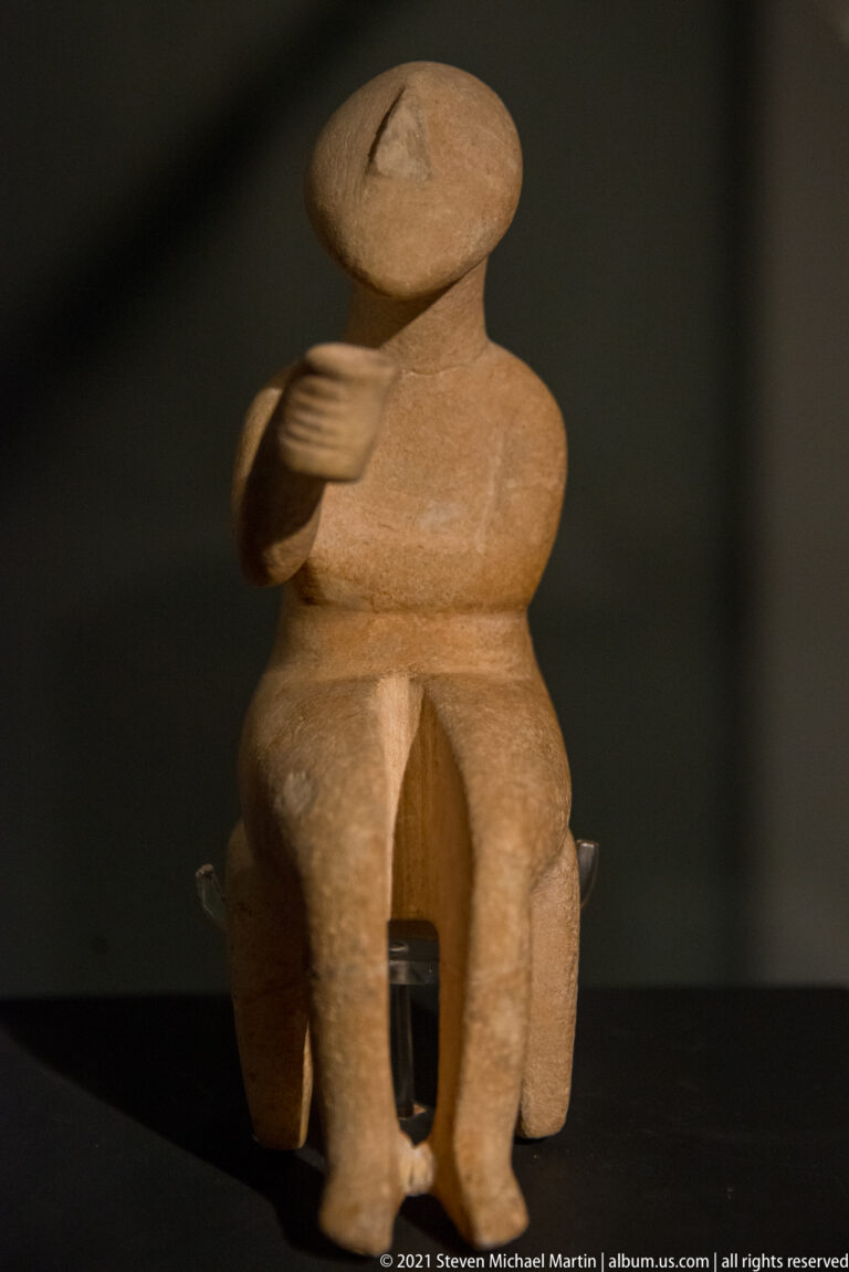 Seated pale stone anthropomorphic figure with oval head, large thighs, separated legs, and one arm raised across the chest.