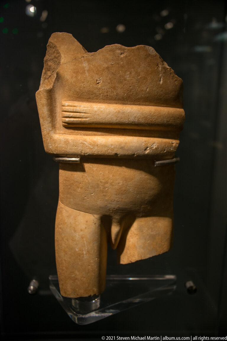 Stone figurine fragment showing crossed forearms over the abdomen, pubic detail, and upper legs with ancient breaks.