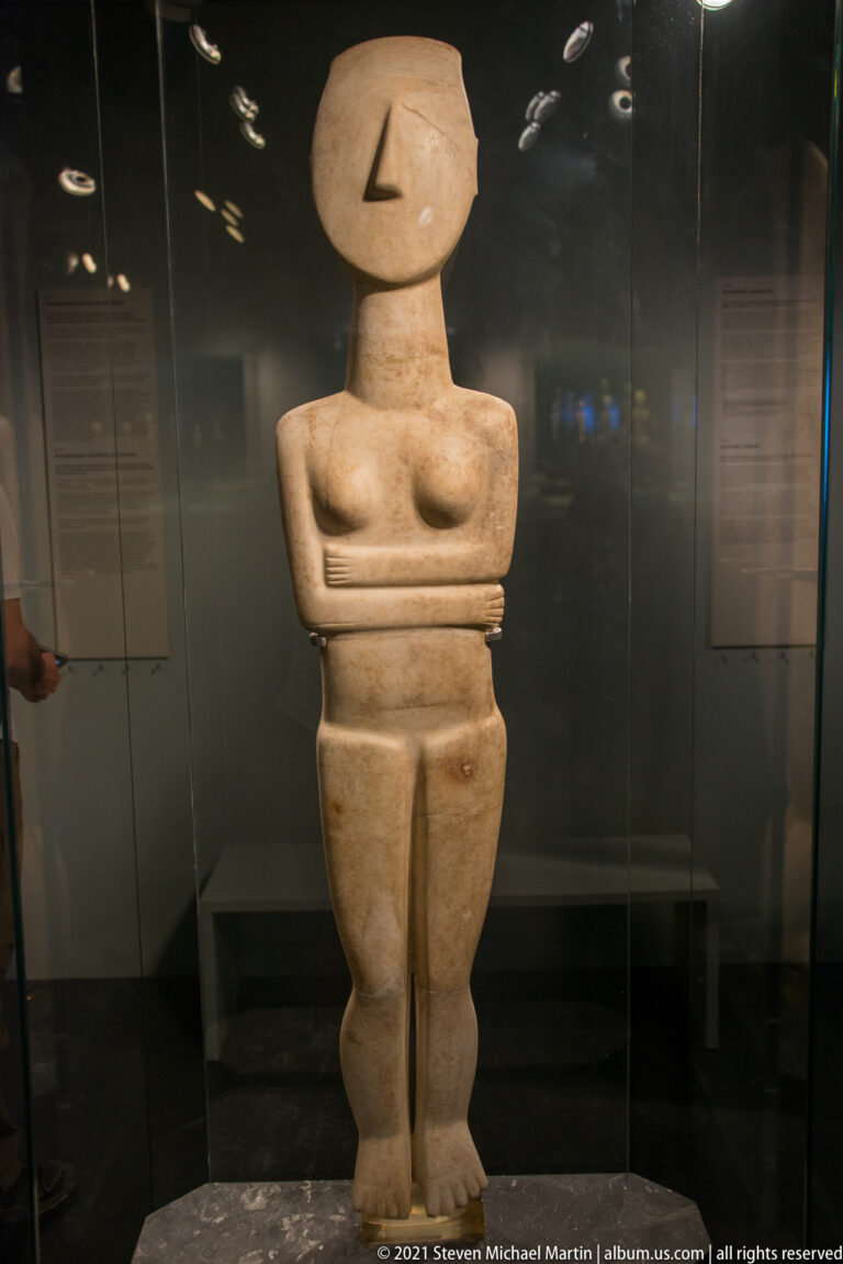 Tall Cycladic marble female figurine with oval head, projecting nose, modeled breasts, folded arms, and long divided legs.