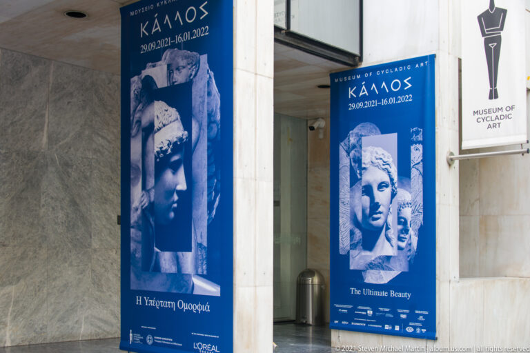 Blue Kallos exhibition banners with sculpted heads beside the glass entrance and vertical Museum of Cycladic Art sign.