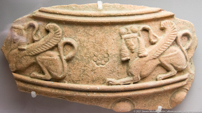 Close view of a coarse ceramic vessel with raised applied human and animal figures arranged around its outer wall.