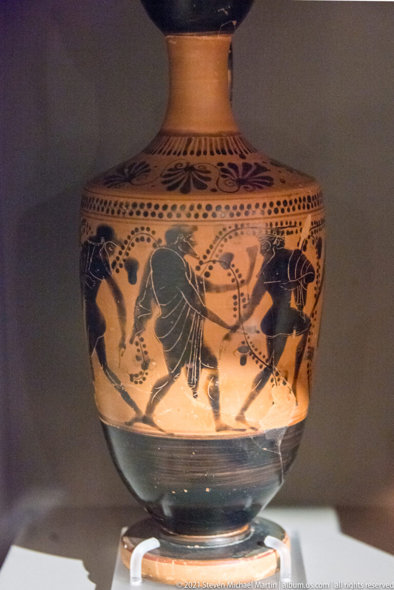 Black-figure Greek ceramic vessel with several dark silhouetted figures and incised details framed by decorative bands.