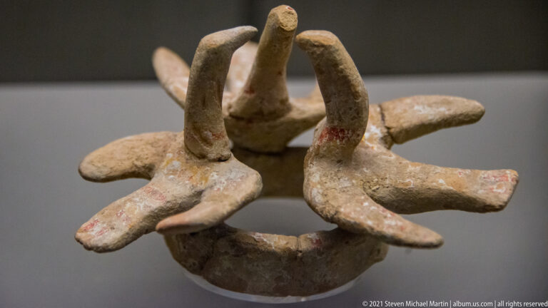 Top view of a pale ceramic ring vessel with multiple radial projections, two tall looped elements, and traces of red pigment.