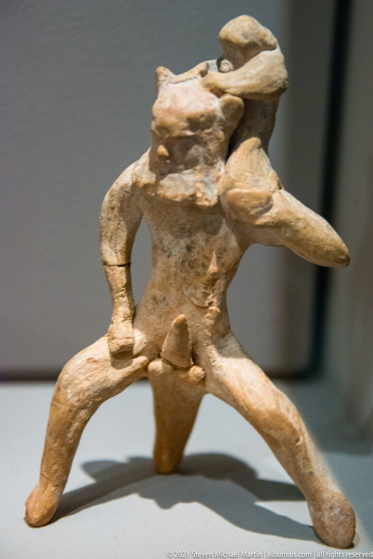 Small weathered terracotta human figure with widely spread legs and asymmetrically extended arms in a dynamic pose.