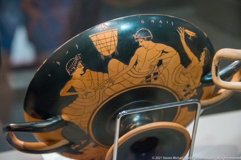 Interior of a red-figure Greek drinking cup with reclining figures, black ground, Greek inscription, handle, and foot visible.