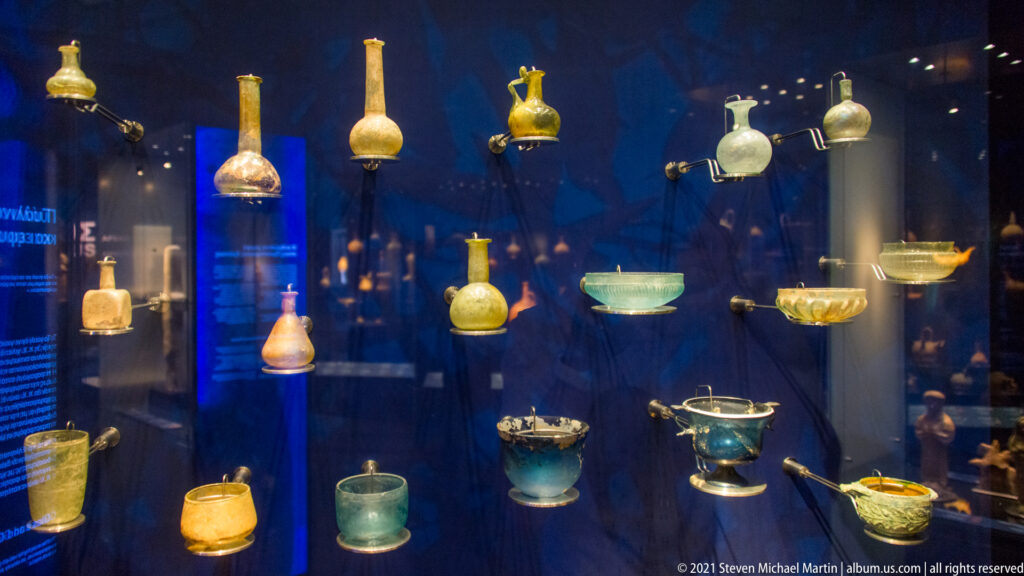 Blue museum case displaying many ancient glass bottles, bowls, cups, and jars.