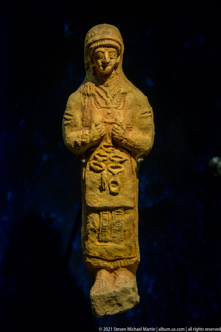 Front-facing terracotta human figurine with folded arms and a broad head covering.