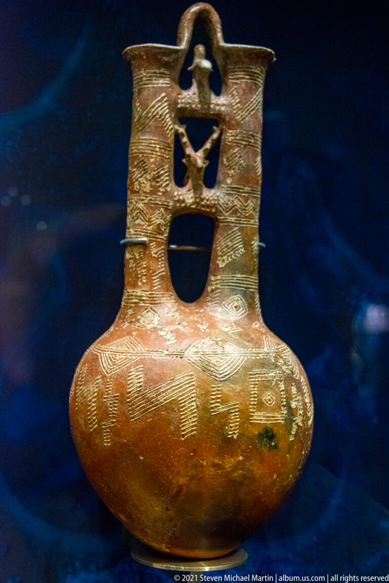 Reddish ceramic vessel with rounded body, incised decoration, and tall pierced neck.