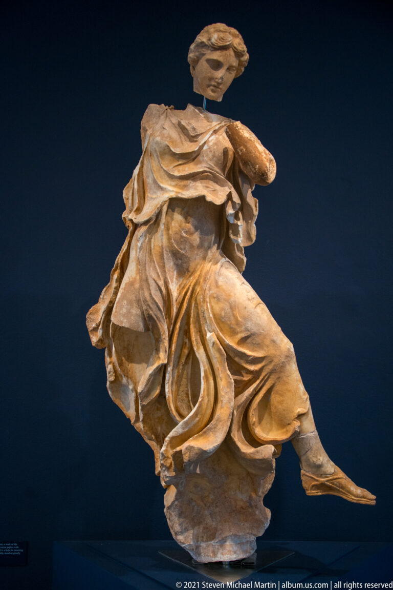 Fragmentary marble female figure in flowing drapery, posed dynamically against a dark museum display background.