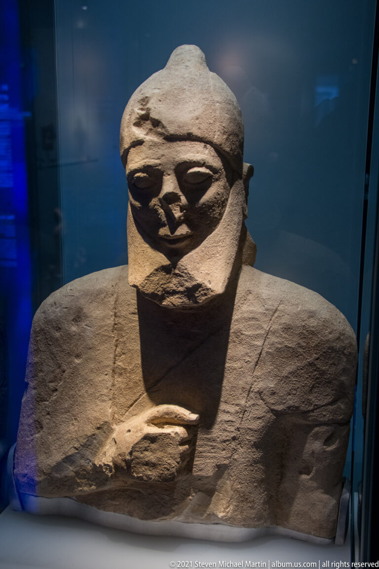 Large frontal stone figure with broad shoulders, carved face, and hands held at the chest.