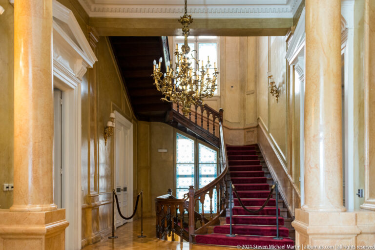 Grand red-carpeted staircase with carved balustrade, columns, tall windows, and chandelier.