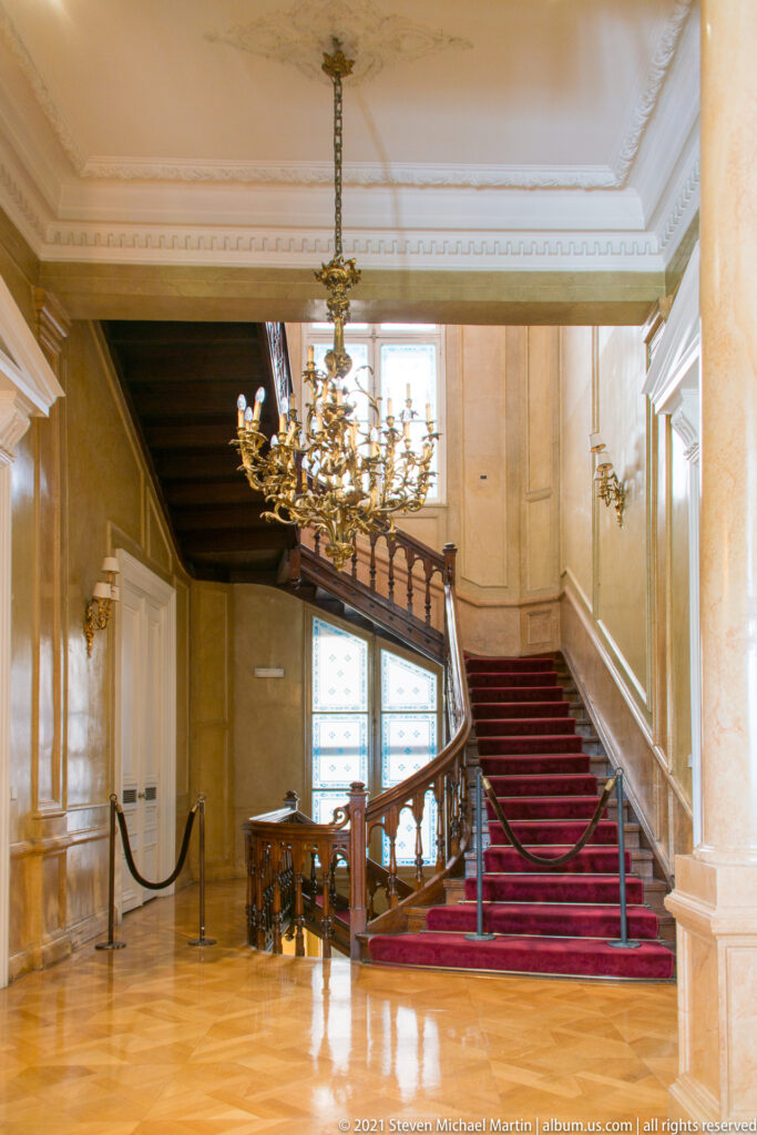 Wide interior view of parquet floor leading to a red-carpeted grand staircase and chandelier.