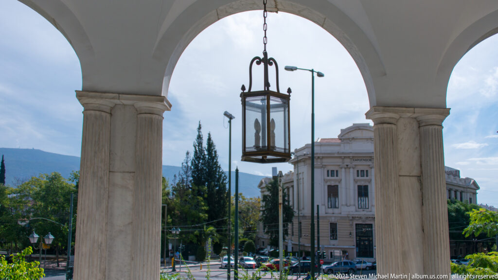 Hanging lantern framed by stone columns and arches, with street and buildings beyond.