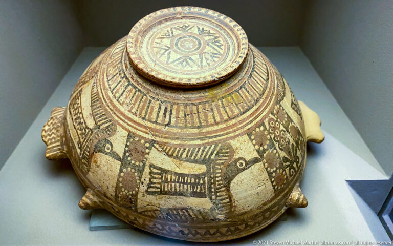 Rounded lidded ceramic vessel with dense geometric painting and small projecting feet or animal forms.
