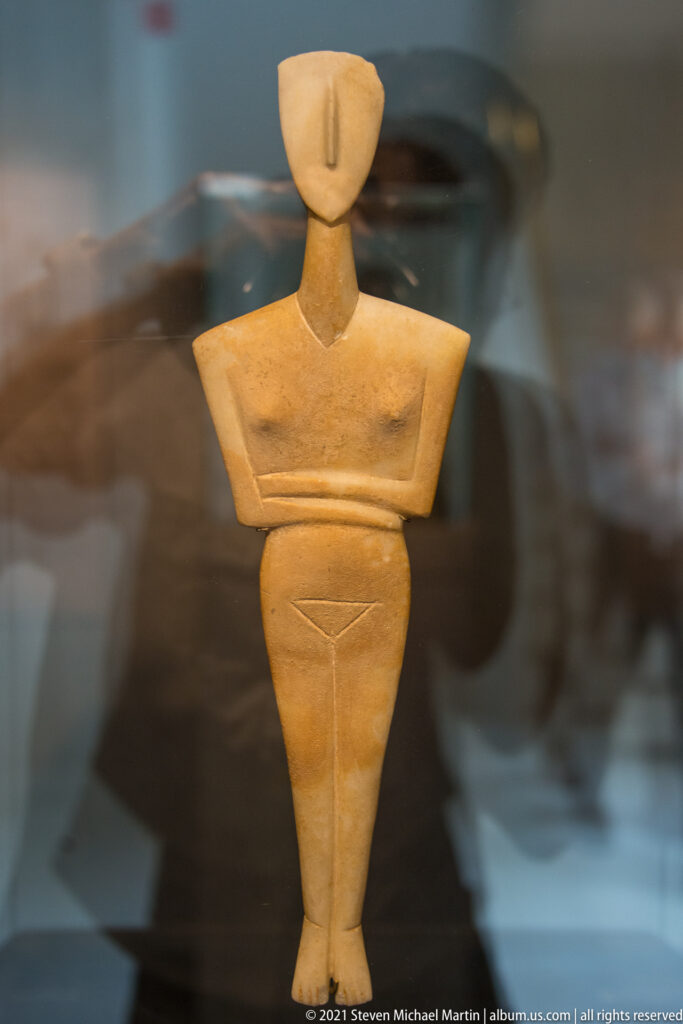 Pale marble Cycladic female figurine with folded arms, long nose, angular shoulders, and simplified legs behind glass.