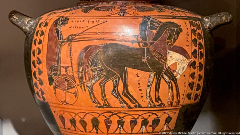 Black-figure ceramic vessel with horse-drawn chariot scene, human figures, decorative borders, and a handle at right.