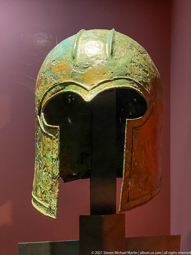 Corinthian bronze helmet dated 475–450 BC with rounded crown, nose-guard, cheek-pieces, and green-brown patina.