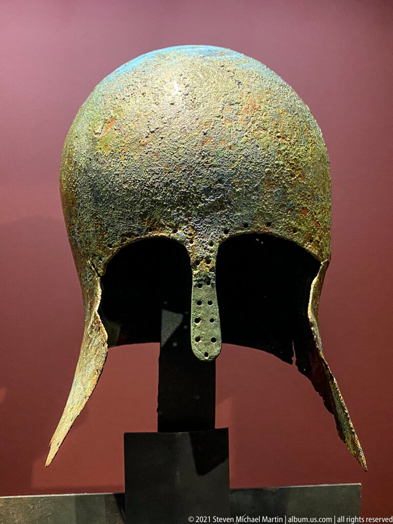 Front view of a Corinthian bronze helmet dated 475–450 BC with eye openings, nasal guard, and long cheek-pieces.