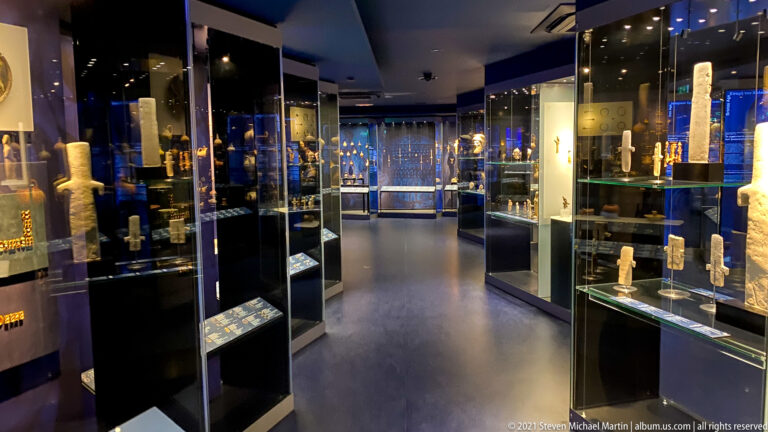 Dark museum gallery lined with illuminated glass cases displaying small ancient sculptures, vessels, and other antiquities.