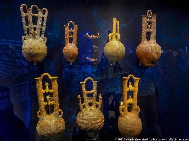 Museum case with several round ceramic vessels bearing unusually tall openwork handles and painted geometric decoration.